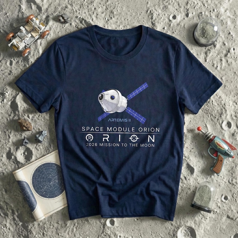 May include: Navy blue t-shirt featuring a graphic of the Artemis II Space Module Orion with text that reads "2026 Mission to the Moon." The shirt is surrounded by toy space-themed items, including a toy rocket and a lunar rover.
