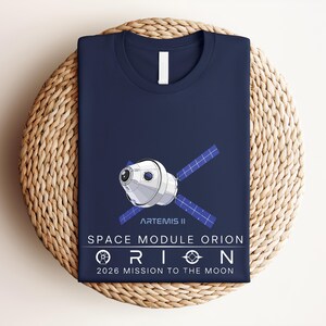 May include: Navy blue t-shirt featuring a graphic of the Artemis II Space Module Orion. The design includes the text "SPACE MODULE ORION" and "2026 MISSION TO THE MOON". The shirt is folded and placed on a woven, circular surface.