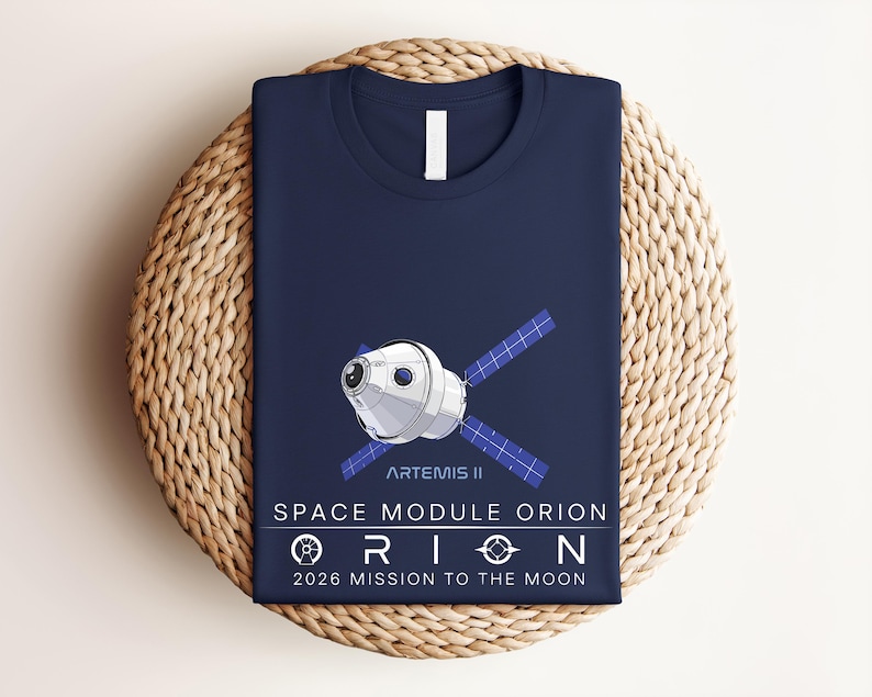 May include: Navy blue t-shirt featuring a graphic of the Artemis II Space Module Orion. The design includes the text "SPACE MODULE ORION" and "2026 MISSION TO THE MOON". The shirt is folded and placed on a woven, circular surface.
