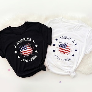 Może przedstawiać: Dwie koszulki, czarna i biała, z patriotycznym wzorem. Każda ma postarzaną grafikę amerykańskiej flagi, napis "AMERICA" i daty "1776 - 2026". Koszulki są złożone.