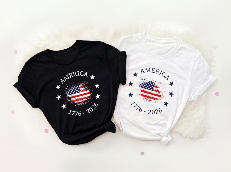 Może przedstawiać: Dwie koszulki, czarna i biała, z patriotycznym wzorem. Każda ma postarzaną grafikę amerykańskiej flagi, napis "AMERICA" i daty "1776 - 2026". Koszulki są złożone.