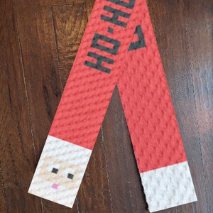 May include: A red Christmas scarf with the text "HO-HO" in black. The scarf features a pixelated Santa Claus face at one end and white trim at the other. The scarf is made of a textured material.