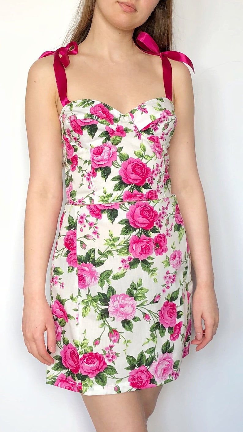 May include: A white dress with a pink floral print and pink ribbon straps. The dress has a fitted bodice and a flared skirt.