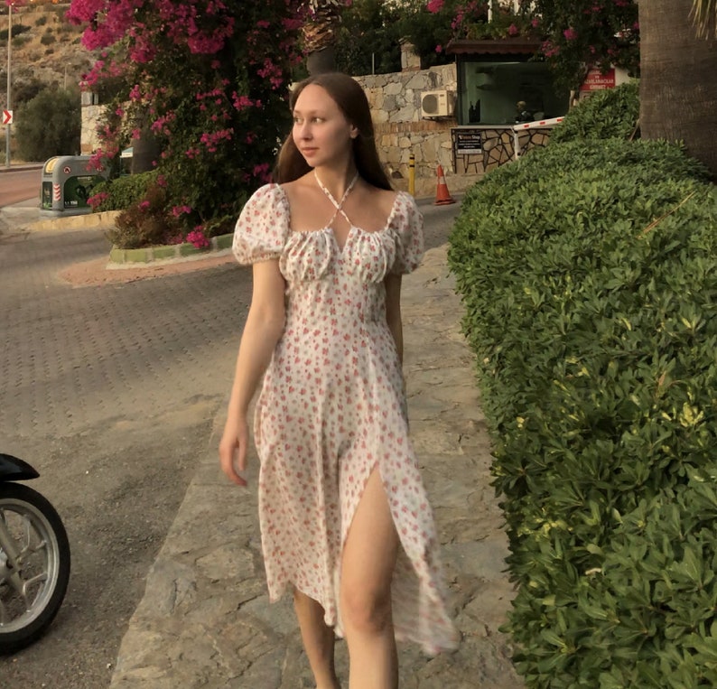May include: A white floral print dress with a halter neckline and puff sleeves. The dress features a thigh-high slit and a midi length. The background includes greenery, a road, and a building.