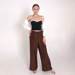 May include: A woman wearing a black strapless top with a white shirt tied around her waist and brown wide-leg pants.
