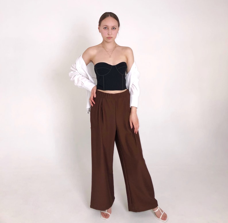 May include: A woman wearing a black strapless top with a white shirt tied around her waist and brown wide-leg pants.