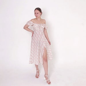 May include: Off-the-shoulder midi dress in white with a delicate floral pattern in red. The dress features a thigh-high slit and is paired with white heeled sandals. The style is feminine and suitable for warm weather.