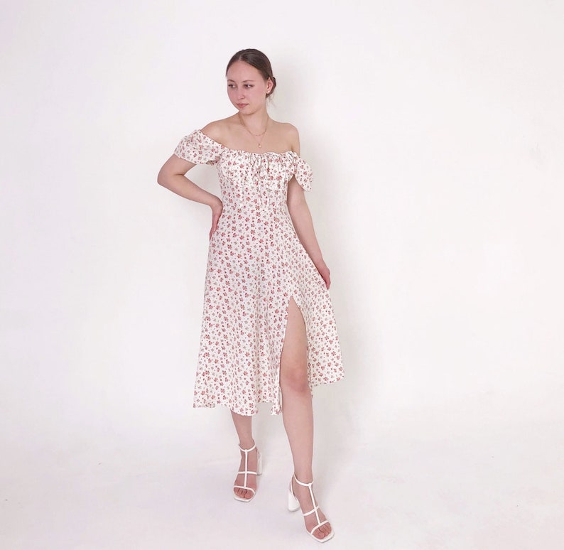 May include: Off-the-shoulder midi dress in white with a delicate floral pattern in red. The dress features a thigh-high slit and is paired with white heeled sandals. The style is feminine and suitable for warm weather.