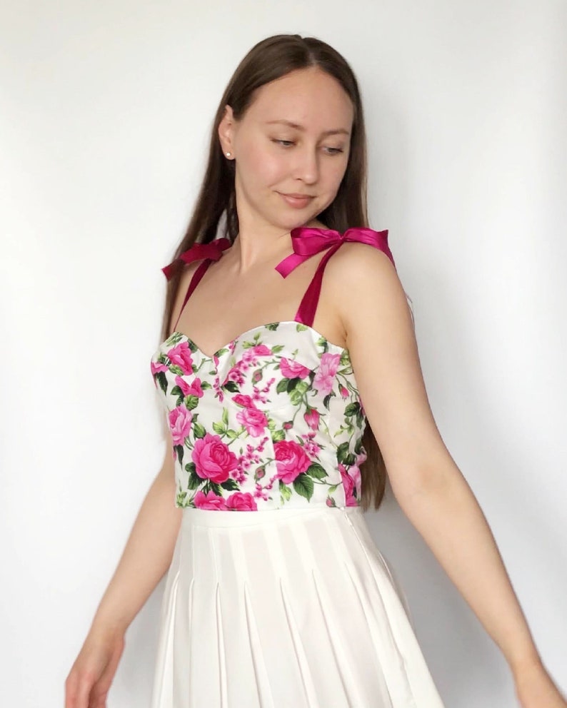 May include: A white crop top with a floral print featuring pink roses and green leaves. The top has pink satin straps that tie into bows at the shoulders.