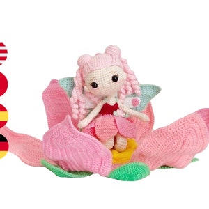 May include: A pink crocheted fairy doll sitting in a pink crocheted flower. The fairy has pink hair, a red dress, and green wings.