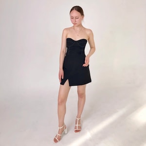 May include: A black strapless mini dress with a fitted bodice and a side slit. The dress has a seam detail around the bodice and skirt. The model is wearing white heeled sandals.