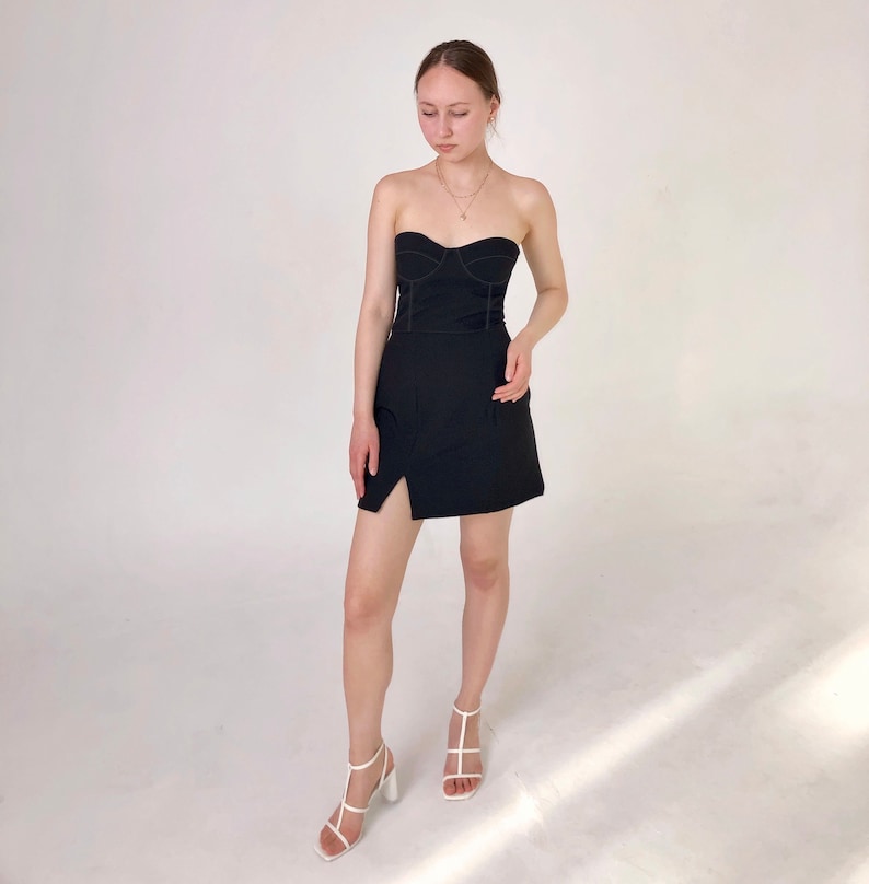 May include: A black strapless mini dress with a fitted bodice and a side slit. The dress has a seam detail around the bodice and skirt. The model is wearing white heeled sandals.