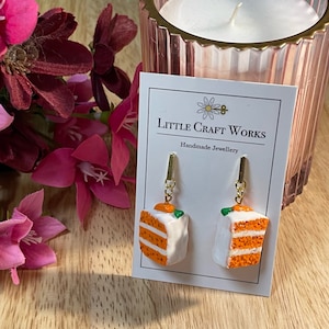 May include: A pair of handmade earrings featuring miniature carrot cake slices. Each earring has a gold-colored hook and a small, detailed cake slice with orange layers, white frosting, and a tiny carrot decoration. The card reads "Little Craft Works Handmade Jewellery."