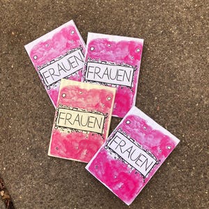 May include: Four small, handmade cards with the word "FRAUEN" in a black outlined font. The cards have a pink watercolor background with white accents. The cards are arranged on a textured, gray surface.