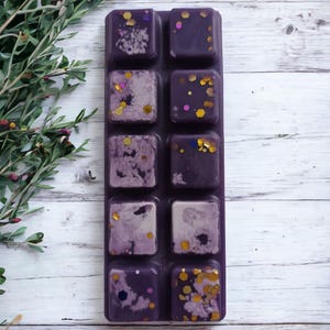 May include: A dark purple wax melt bar with ten individual squares, each decorated with gold and iridescent glitter. Some squares have a marbled, light purple design. The bar is on a white wooden surface with green foliage.
