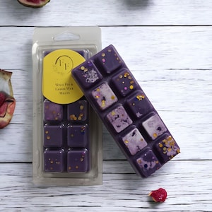 May include: Purple wax melts in a clear plastic packaging with a label that reads "Wild Fig & Cassis Wax Melts". The packaging is on a white wooden surface with fig slices scattered around it.