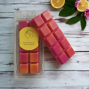 May include: Two bars of pink and orange soy wax melts, one in a clear plastic container and one loose. The container has a yellow label that reads "Pink Lemonade Soy Wax Melts". The background includes lemon slices and flowers.