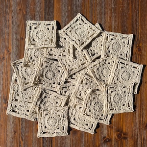 Peut inclure: Un ensemble de napperons carrés au crochet de couleur crème disposés sur une surface en bois. Chaque napperon présente un motif floral central entouré d'une bordure ajourée. Les napperons sont de tailles variables et sont faits d'un fil doux et texturé.