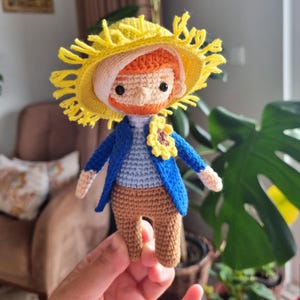 Könnte beinhalten: Eine gehäkelte Puppe mit einem gelben Strohhut, einer blauen Jacke und braunen Hosen. Die Puppe hat einen roten Bart und eine Sonnenblume auf der Brust.
