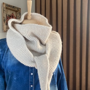 May include: A cream-colored knitted cowl neck scarf with a long, flowing end. The scarf is draped over a denim shirt.