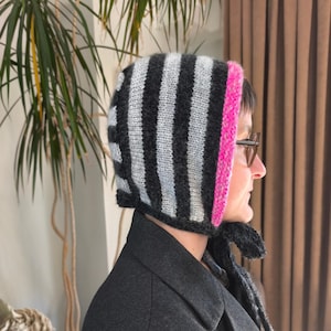 Könnte beinhalten: Eine gestrickte Mütze mit vertikalen Streifen in Grau, Schwarz und Hellblau. Die Mütze hat eine leuchtend rosa Einfassung und Bindebänder unter dem Kinn. Die Mütze wird mit einem dunkelgrauen Mantel getragen.