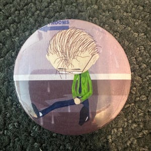 May include: A round button with a cartoon illustration of a person with glasses and a green shirt standing in the rain. The person is holding onto a sign that says "Rooms".