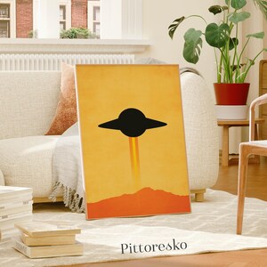 May include: A framed art print featuring a black UFO design against a yellow and orange background. The artwork includes a mountain silhouette and the text "Pittoresko" at the bottom. The print is displayed in a living room setting.