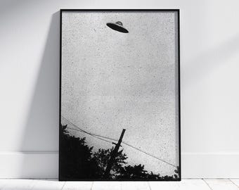 May include: Black and white photograph of a flying saucer in the sky above a power line and trees. The image is framed in a black frame.