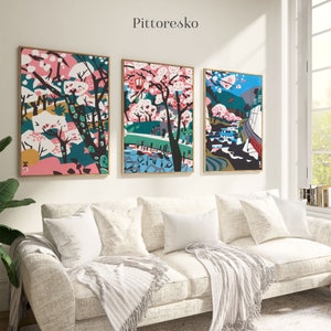 Könnte beinhalten: Drei gerahmte Kunstdrucke mit japanischem Thema. Die Drucke zeigen farbenfrohe Illustrationen von Kirschblüten, Bäumen und Landschaften. Die Drucke hängen an einer weißen Wand über einem hellen Sofa.