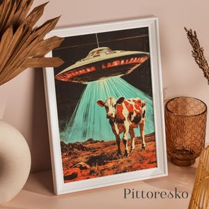 May include: A framed art print depicts a cow being beamed up by a UFO. The artwork features a red and white cow standing on a reddish-brown landscape under a teal beam of light. The UFO is silver and red, set against a dark starry sky.