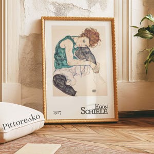 Può includere: Stampa incorniciata di un'opera d'arte di Egon Schiele. L'opera raffigura una figura seduta con i capelli rossi, che indossa un top verde e leggings neri. La cornice è dorata e la stampa è datata "1917" e firmata "Egon Schiele".