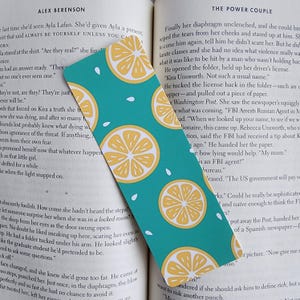 May include: A teal blue bookmark with a repeating pattern of yellow lemons on a turquoise background. The bookmark has white seed-like shapes scattered around the lemons. The bookmark is partially covering a book page with text about a Russian agent.