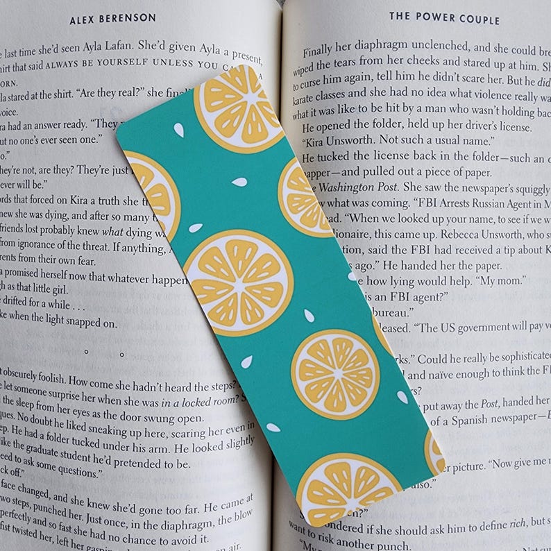 May include: A teal blue bookmark with a repeating pattern of yellow lemons on a turquoise background. The bookmark has white seed-like shapes scattered around the lemons. The bookmark is partially covering a book page with text about a Russian agent.