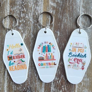 May include: Three white keychains with silver key rings. Each keychain has a different design with text and illustrations related to reading. One says "Hello I'd rather be reading", another says "I have no shelf control", and the last says "In my bookish era".