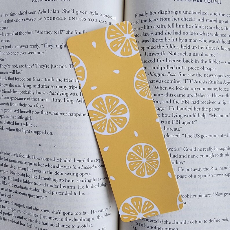 May include: A yellow bookmark with a repeating pattern of white lemons. The bookmark is made of thick paper and has a slightly textured surface.