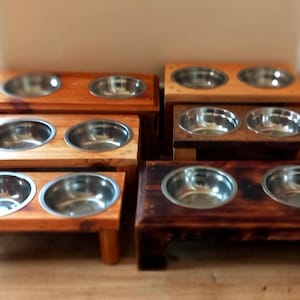 Könnte beinhalten: Eine Sammlung von Futternäpfen aus Holz mit Edelstahleinsätzen. Die Näpfe sind in verschiedenen Brauntönen, von hell bis dunkel, und stehen auf kleinen Holzständern. Die Anordnung umfasst Einzel- und Doppel-Napf-Designs.