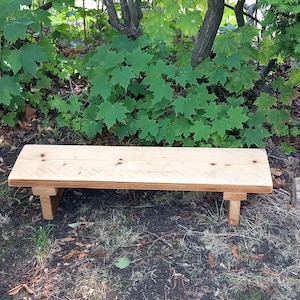 Op de afbeelding: Een kleine, rechthoekige houten bank met vier poten. De bank is gemaakt van lichtgekleurd hout en staat buiten in een natuurlijke omgeving met groene bladeren op de achtergrond. De bank is ongeveer 122 cm lang.