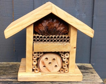May include: A wooden bee house with a pitched roof, featuring multiple compartments for nesting. The design includes a pine cone, bamboo tubes, and a wooden disc with holes. The natural wood tones create a rustic aesthetic.