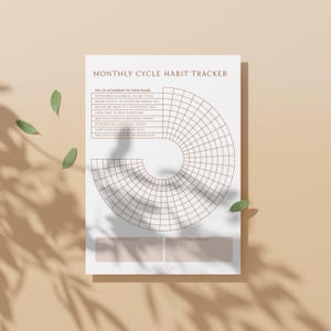 May include: A habit tracker with the text "MONTHLY CYCLE HABIT TRACKER". The design features a circular grid for tracking habits, with prompts for self-care. The paper is white, set against a light brown background, with green leaves.