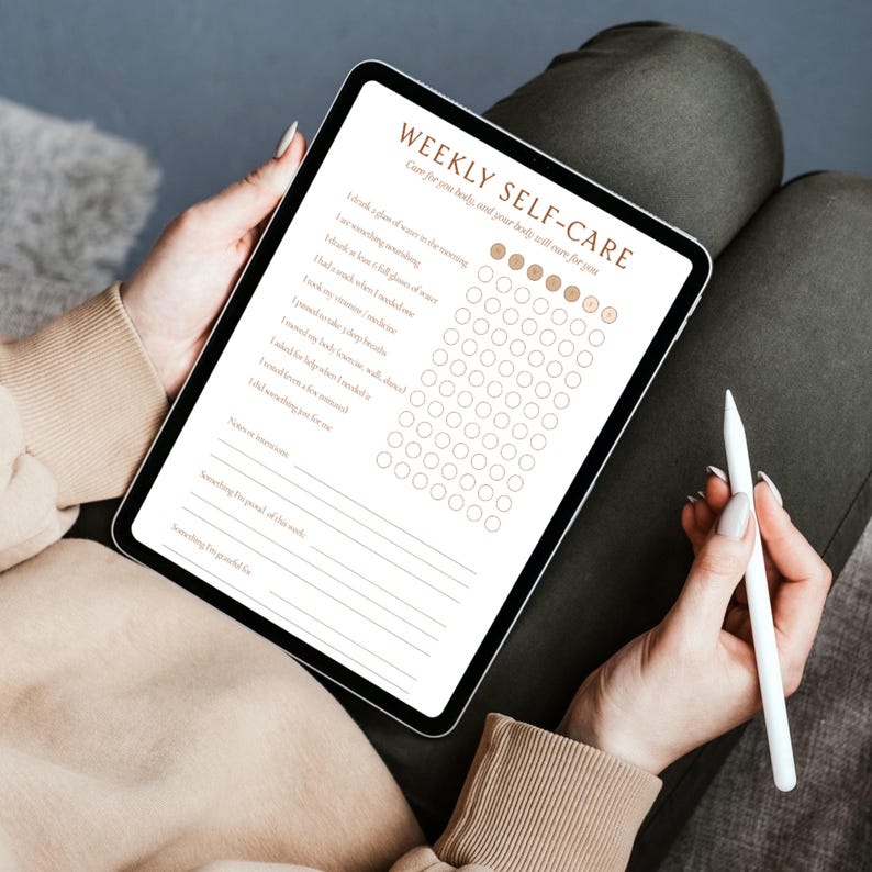 May include: A digital tablet displaying a "WEEKLY SELF-CARE" checklist. The screen shows a list of self-care activities with checkboxes. A white stylus rests in the hand of the person holding the tablet. The tablet has a black border.