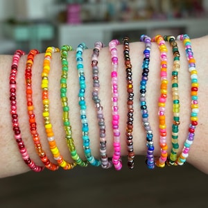 Può includere: Una collezione di undici braccialetti sottili realizzati con piccole perline di vetro rotonde e multicolori. I braccialetti presentano varie tavolozze di colori tra cui rosso, arancione, giallo, verde, blu, rosa, viola e toni metallici in file.