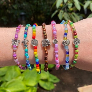 Puede incluir: Una colección de pulseras de cuentas en varios colores, incluyendo morado, azul, verde y arcoíris. Cada pulsera presenta un dije diferente, como una mariposa, una tortuga, un signo de paz, un elefante, una flor, una luna creciente y un árbol de la vida.