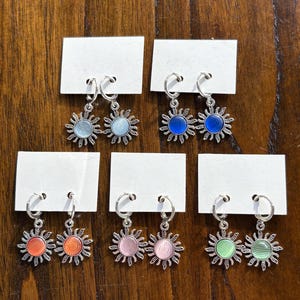 May include: A collection of silver-toned hoop earrings, each featuring a sunburst design with a colorful center. The earrings are displayed on white cards against a wooden background. The sunburst centers are in shades of blue, orange, pink, and green.