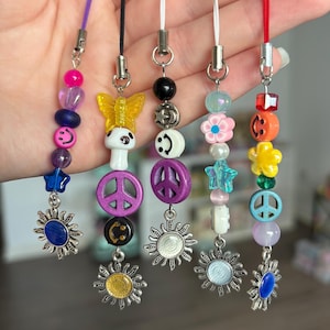 May include: A collection of beaded phone charms in various colors. Each charm features a different design, including smiley faces, peace signs, stars, and flowers. The charms are attached to colorful cords.
