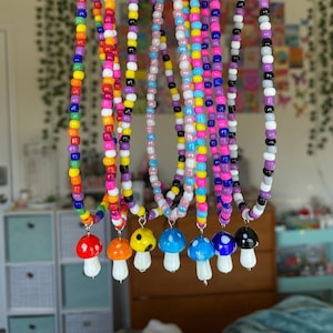 May include: A collection of beaded necklaces with mushroom pendants. The necklaces feature colorful beads in various patterns, with mushroom charms in red, orange, yellow, blue, and black. Each mushroom has a white base. The necklaces are hanging against a blurred background.
