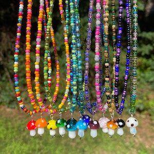 May include: A selection of beaded necklaces with mushroom pendants. The necklaces display a range of bead colours, including orange, green, blue, and purple. Each necklace has a different coloured mushroom pendant with a white stem.