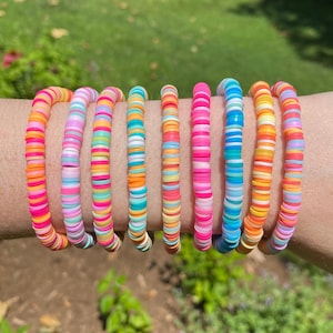 May include: A collection of colorful, flat disc bracelets in various shades of pink, orange, yellow, blue, and green. The bracelets are stacked on top of each other, showcasing a vibrant, rainbow-like effect. The bracelets are made of small, flat beads.