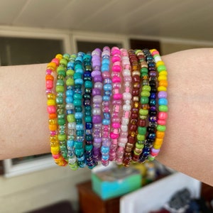 Puede incluir: Una colección de pulseras de cuentas coloridas en varios tonos, incluyendo rosa, verde, azul y amarillo. Las pulseras están apiladas en una muñeca, mostrando la variedad de colores y diseños. Cada pulsera está hecha de pequeñas cuentas redondas.