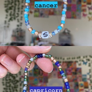 May include: Two beaded bracelets, one blue and gold with "cancer" text, the other blue, green, and gold with "capricorn" text. Each bracelet has a zodiac symbol bead. The bracelets are held up.