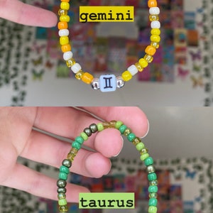 May include: Two beaded bracelets. The top bracelet is yellow, orange, and white with a Gemini symbol. The bottom bracelet is green with a Taurus symbol. Both bracelets are on a white background.